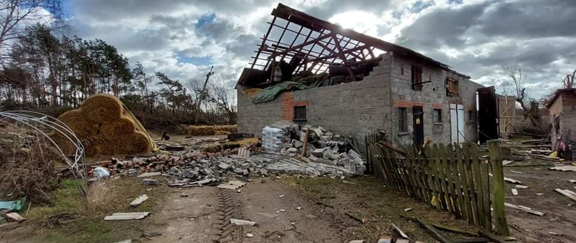 Zdjęcie przedstawia częściowo uszkodzony dach budynku gospodarczego oraz zniszczony szczyt z cegły. Cegły leżą przed budynkiem 