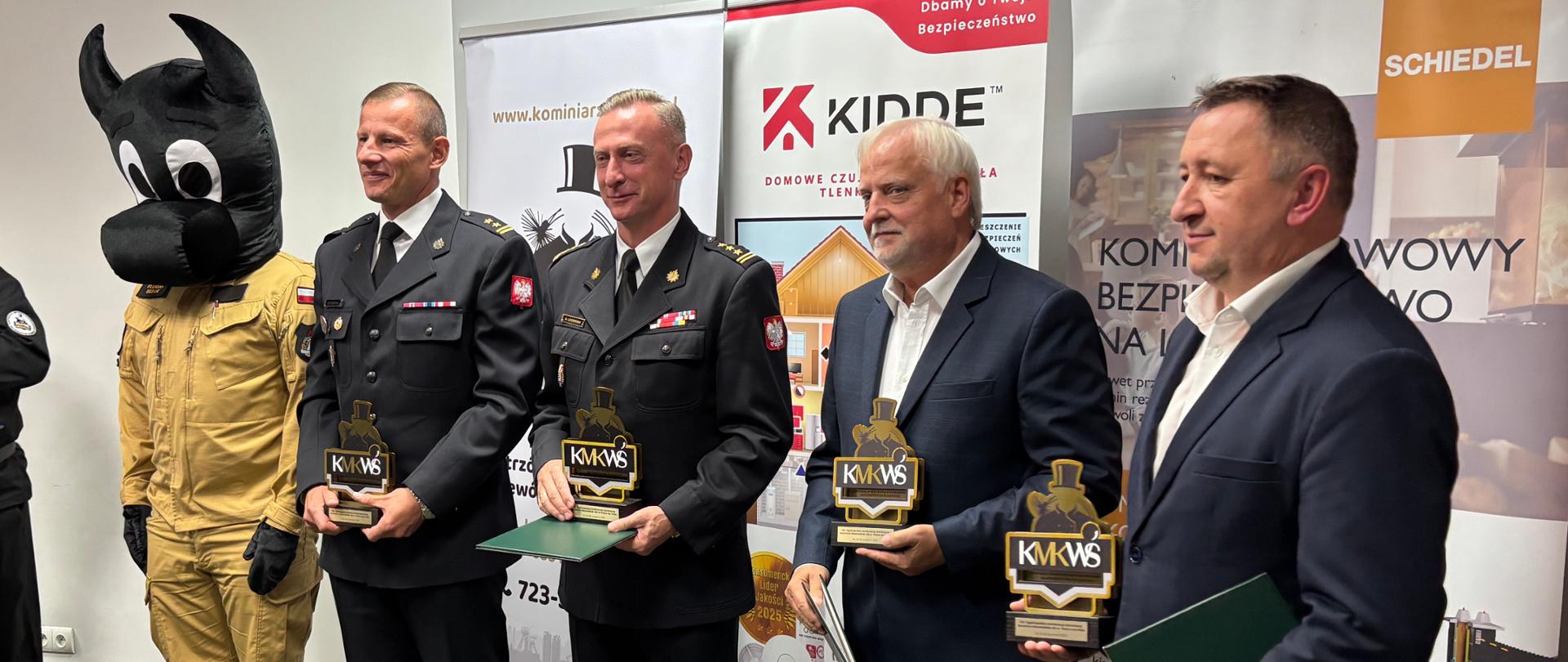 Zdjęcie przedstawia grupę mężczyzn podczas konferencji kominiarzy. W środku, pięciu mężczyzn stoi obok siebie, uśmiechając się do aparatu. Dwóch z nich, ubranych w mundury strażackie, trzyma w rękach pamiątkowe statuetki i dyplomy. Obok nich stoi dwóch mężczyzn w garniturach, również ze statuetkami. Z lewej strony grupy widać postać przebraną w strój beboka -floriana. W tle, za grupą, znajdują się banery reklamowe z logotypami.