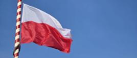 Zdjęcie przedstawia łopoczącą na wietrze flagę Rzeczypospolitej Polskiej podczas podnoszenia na maszt.