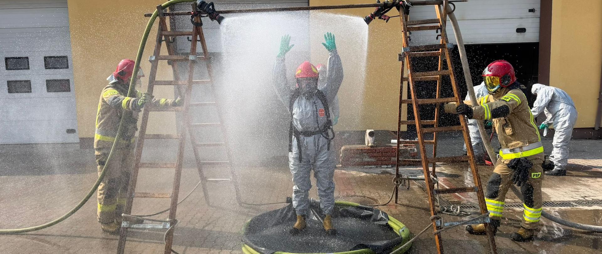 Szkolenie doskonalące z zakresu ratownictwa chemicznego i ekologicznego - strażacy podczas ćwiczeń, w trakcie procesu dekontaminacji. Strażak w basenie, miedzy dwiema drabinami, z których tryska woda. Drabiny trzymane przez strażaków, w tle bramy garażowe Ośrodka Szkolenia