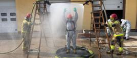 Szkolenie doskonalące z zakresu ratownictwa chemicznego i ekologicznego - strażacy podczas ćwiczeń, w trakcie procesu dekontaminacji. Strażak w basenie, miedzy dwiema drabinami, z których tryska woda. Drabiny trzymane przez strażaków, w tle bramy garażowe Ośrodka Szkolenia