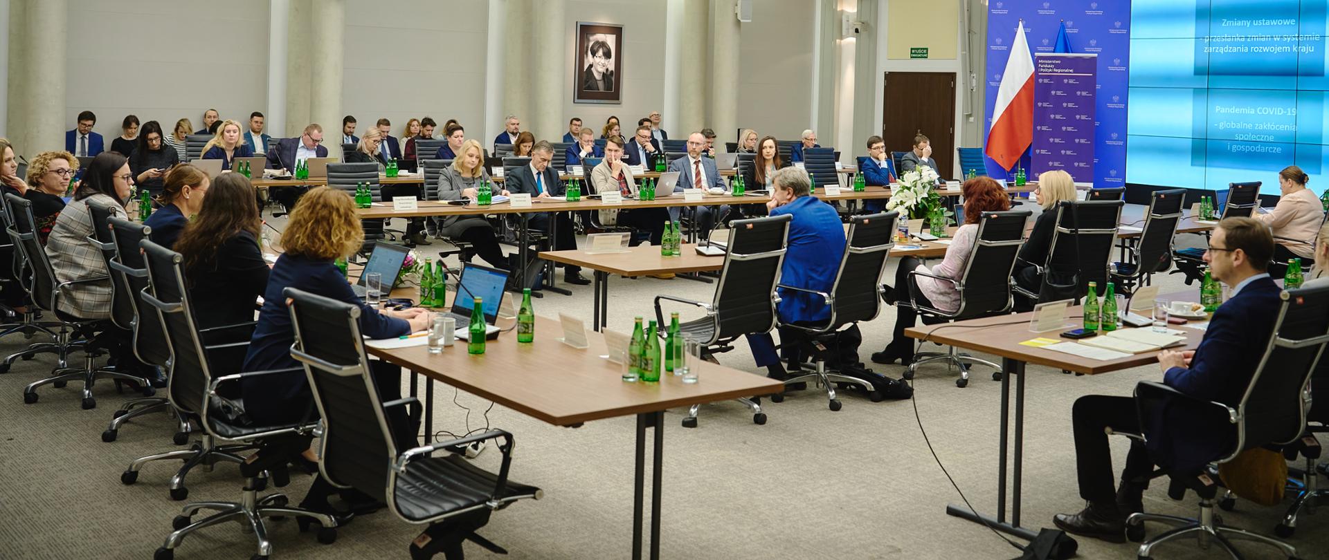 Widok sali konferencyjnej. Trwa spotkanie. Osoby siedzą przy stołach.