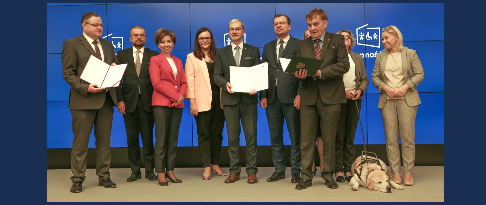 Dziesięć osób na scenie, w środku minister Jerzy Kwieciński z podpisaną umową, obok po lewej wiceminister Małgorzata Jarosińska-Jedynak, po prawej stronie leży pies-przewodnik, z tyłu granatowy ekran z logo programu Dostępność Plus