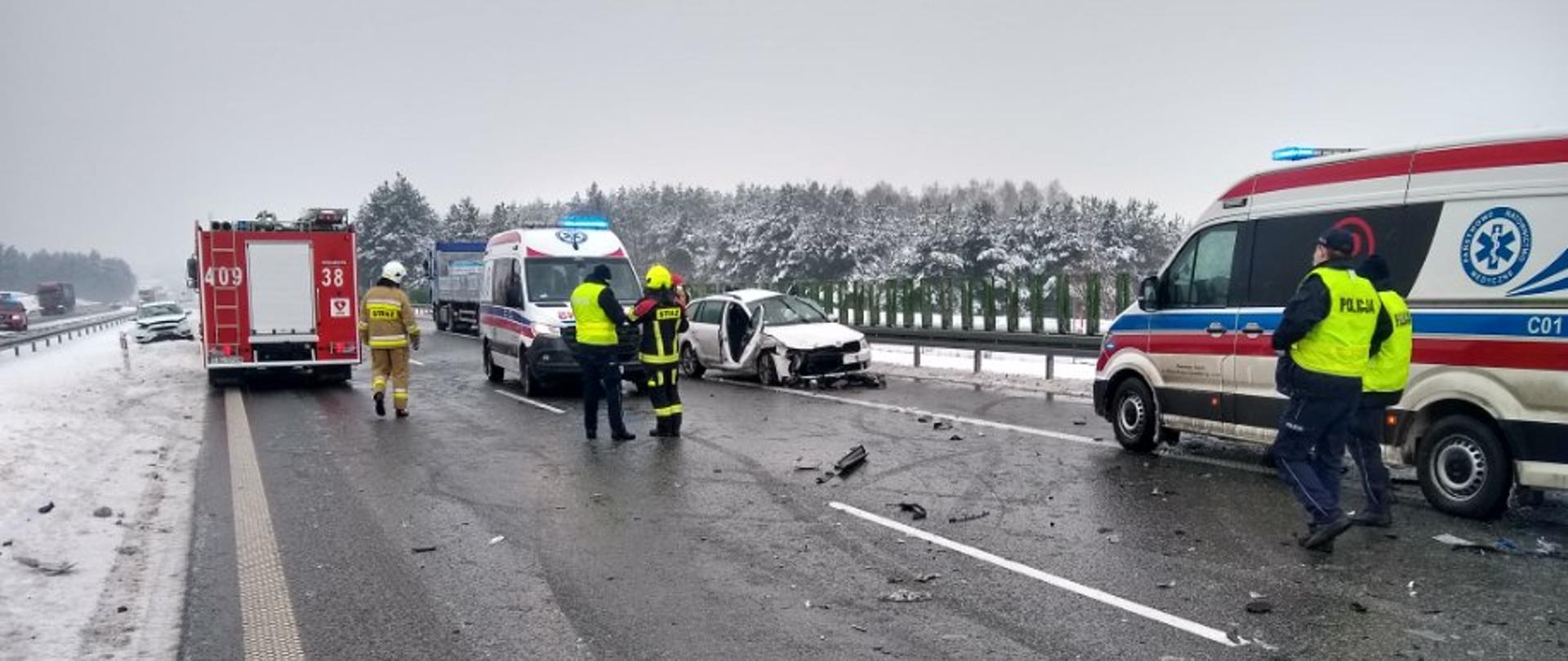 Na zdjęciu widoczne są dwa pasu ruchu autostrady A1. Po prawej stronie widoczna jest karetka pogotowia, przy której znajduje się dwóch policjantów. Karetka ustawiona jest przodem w lewą stronę. Na jej lewym boku widoczne jest logo jednostki ochrony zdrowia oraz fragment numeru bocznego C01. Obok karetki na pasach ruchu widoczne są drobne, wykonane z tworzywa, elementy pojazdów, które zostały oderwane od pojazdów biorących udział w wypadku. Przed karetka znajduje się samochód osobowy zwrócony w prawą stronę. Pojazd ma widoczne uszkodzenia powstałe na skutek wypadku takie jak zniszczona maska, oberwany zderzak, oberwane drzwi pasażera. Obok samochodu osobowego znajduje się druga karetka, zwrócona w tym samym kierunku. Przed karetka stoi strażak i policjant. Dalej z lewej strony, na wysokości karetki widać tył ustawionego samochodu ratowniczo – gaśniczego. Na czerwonym tle pojazdu widoczne są numery operacyjne 409 38. Przy samochodzie widoczny jest strażak. W tle widoczne są samochód osobowy oraz ciężarowy, które również brały udział w wypadku. Po lewej stronie zdjęcia widać pobocze jezdni, pokryte śniegiem. Za barierkami oddzielającymi pasy ruchu widać inne pojazdy. W tle widoczne są barierki ochronne, drzewa pokryte śniegiem oraz ciemno – szare niebo.
