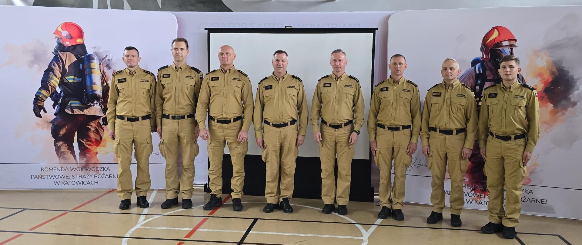 Oficjalne zdjęcie grupowe ośmiu funkcjonariuszy Państwowej Straży Pożarnej w piaskowych mundurach służbowych, stojących w hali sportowej na tle ścianek reklamowych Komendy Wojewódzkiej PSP w Katowicach.