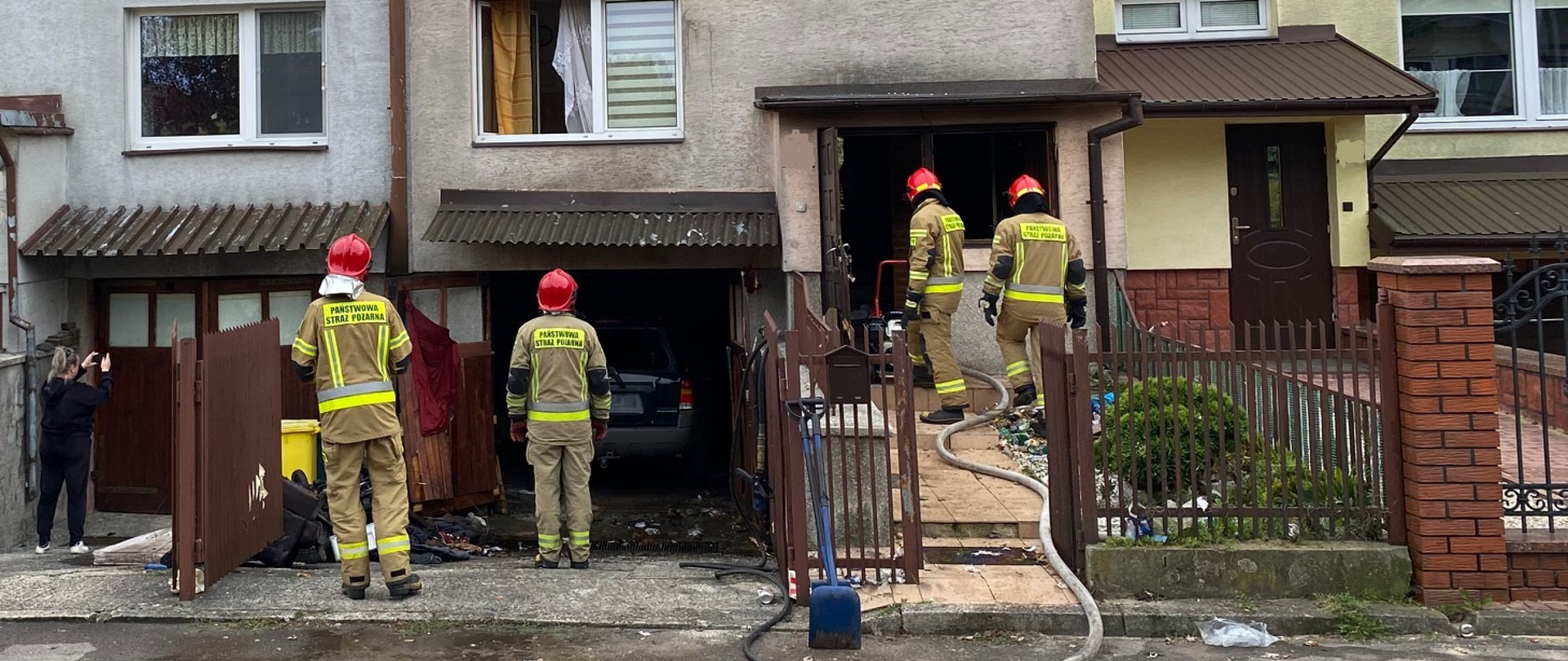 Strażacy przed budynkiem mieszkalnym w którym uszkodzona została brama garażowa i wybite zostały szyby na klatce schodowej. Porozrzucane elementy wyposażenia znajdują się na podjeździe.