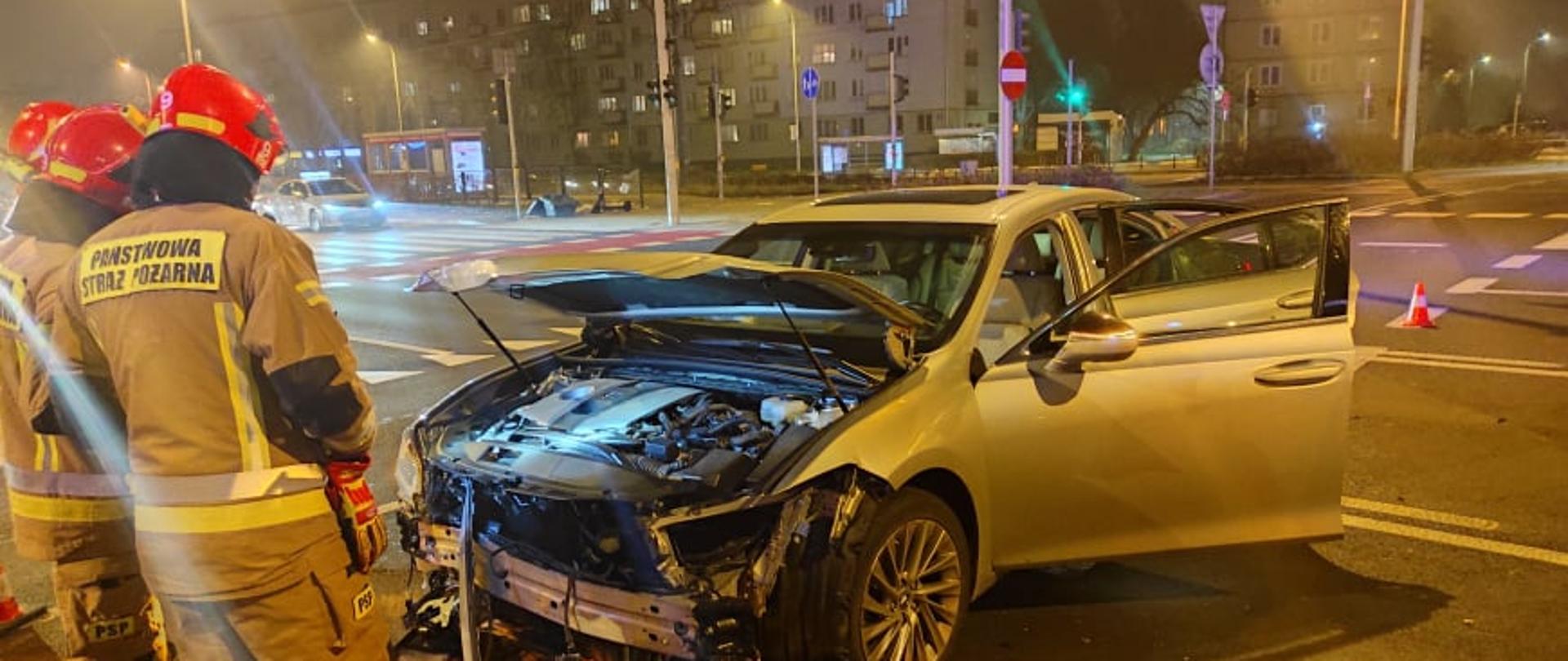 Na zdjęciu widoczny jest rozbity samochód osobowy, ma rozbity przód obok pracujący strażacy.
