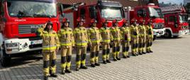 Na zdjęciu widoczni strażacy na tle budynku komendy i samochodów pożarniczych. Po lewej stronie zdjęcia zdjęcia masz z flagą na środku salutujący strażak dowódca zmiany służbowej obok niego po prawej stronie zmiana służbowa w jednym szeregu. Strażacy ubrani w czerwone hełmy i ubrania specjalne koloru piaskowego.