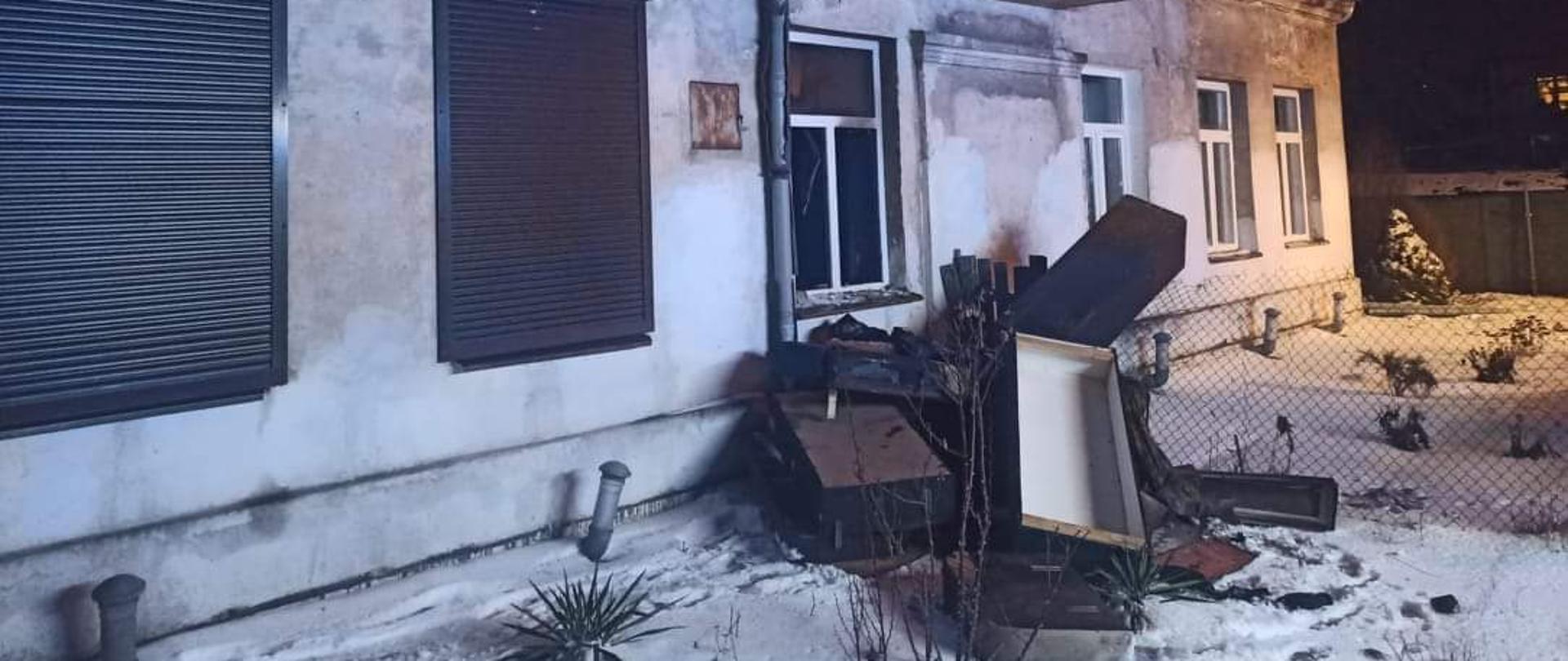 Na zdjęciu widoczny jest budynek mieszkalny, z elewacją koloru białego. Przedstawiona jest dłuższa ściana, w której znajduje się sześć okien – dwa okna po lewej stronie mają zasłonięte żaluzje zewnętrzne koloru brązowego. Przed budynkiem znajduje się sterta mebli – zostały wyniesione z mieszkania w którym był pożar. Zdjęcie wykonano po zmroku, widoczny jest śniegu na trawniku. Przed budynkiem, z gruntu wystaje pięć rur. W poddaszowej części budynku, znajduje się oszklony i zadaszony balkon, obok widoczne jest jedno okno. Na dachu pokrytym blachą znajduje się śnieg.