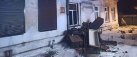 Na zdjęciu widoczny jest budynek mieszkalny, z elewacją koloru białego. Przedstawiona jest dłuższa ściana, w której znajduje się sześć okien – dwa okna po lewej stronie mają zasłonięte żaluzje zewnętrzne koloru brązowego. Przed budynkiem znajduje się sterta mebli – zostały wyniesione z mieszkania w którym był pożar. Zdjęcie wykonano po zmroku, widoczny jest śniegu na trawniku. Przed budynkiem, z gruntu wystaje pięć rur. W poddaszowej części budynku, znajduje się oszklony i zadaszony balkon, obok widoczne jest jedno okno. Na dachu pokrytym blachą znajduje się śnieg.
