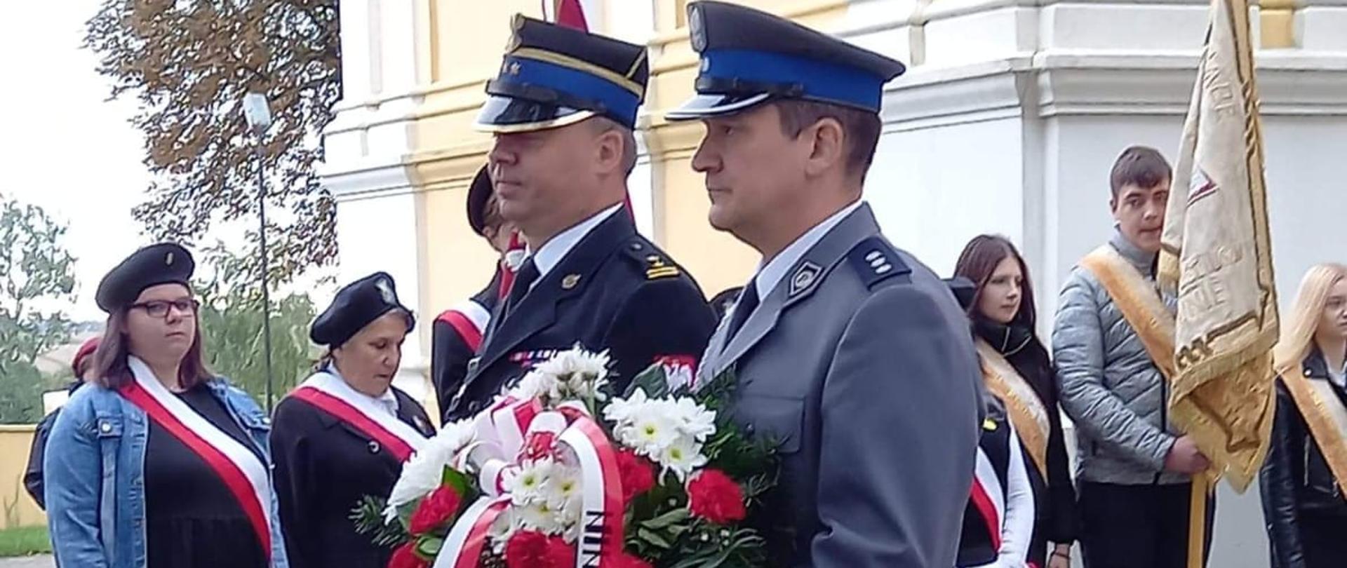 Zdjęcie przedstawia zastępcę komendanta miejskiego oraz policjanta, którzy ubrani są w mundury wyjściowe. Policjant trzyma w ręku kwiaty, które zostaną złożone pod tablicą pamiątkową Sybiraków. Z tytuł widać poczty sztandarowe. 