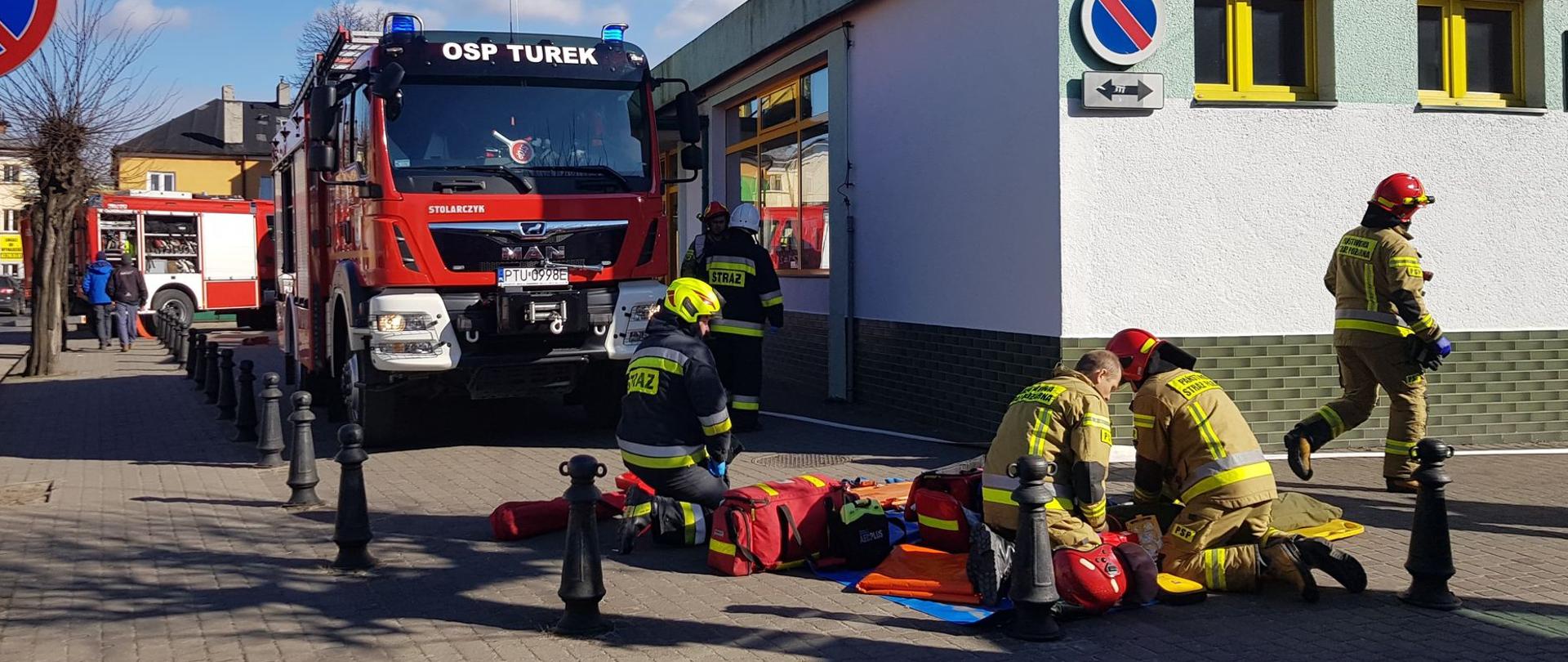 Pojazdy pożarnicze przed budynkiem kina oraz ratownicy w trakcie przygotowywania pola medycznego.
