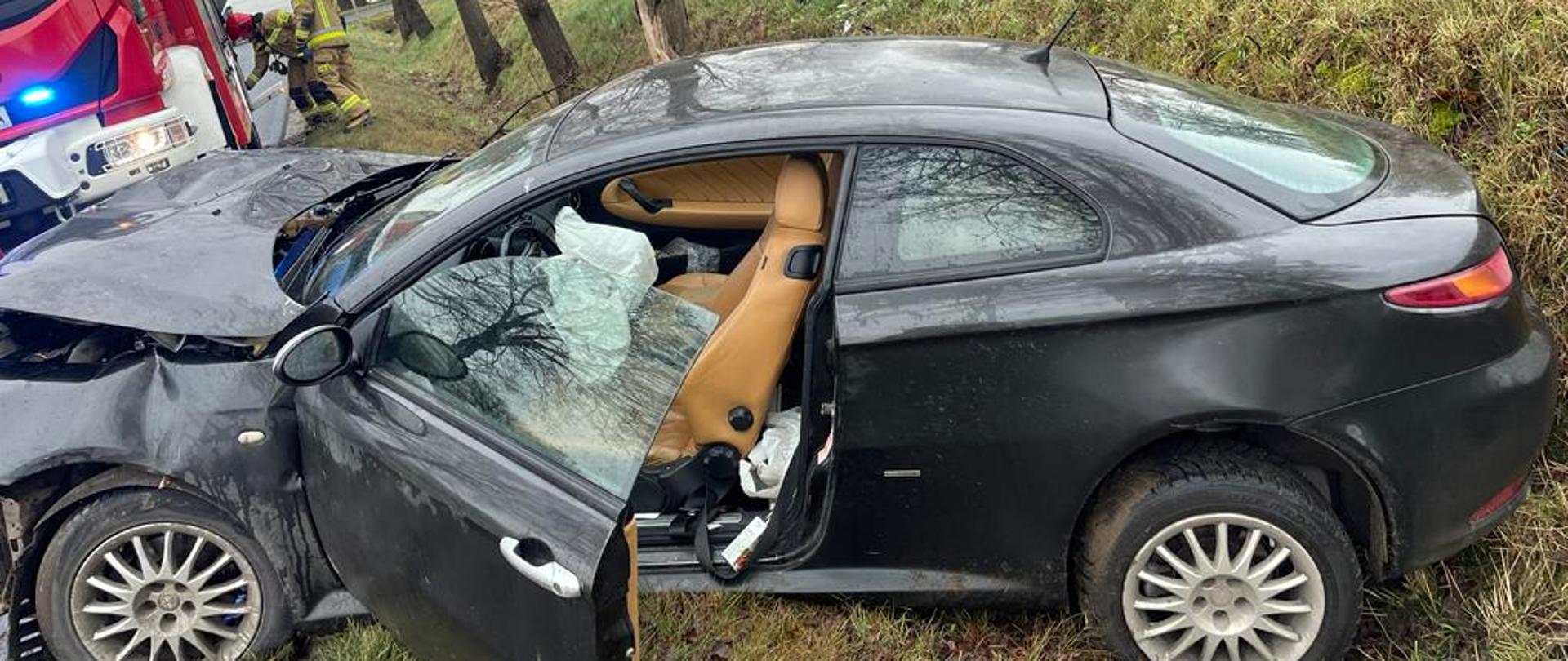 Rozbity samochód osobowy po uderzeniu w drzewo. W tle samochód strażacki i 2 strażaków