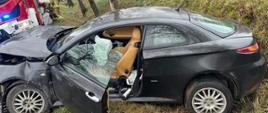 Rozbity samochód osobowy po uderzeniu w drzewo. W tle samochód strażacki i 2 strażaków