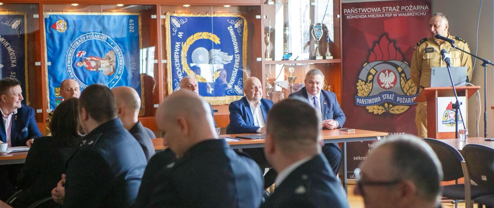Szeroki kadr sali konferencyjnej. W tle, przy stole prezydialnym, siedzą przedstawiciele władz i służb. Po prawej stronie, przy mównicy z logo Państwowej Straży Pożarnej, stoi strażak wygłaszający przemówienie. Ściana za nim ozdobiona jest gablotami ze sztandarami oraz trofeami. Na pierwszym planie widoczne są sylwetki uczestników narady siedzących tyłem do obiektywu, ułożone w rzędach.