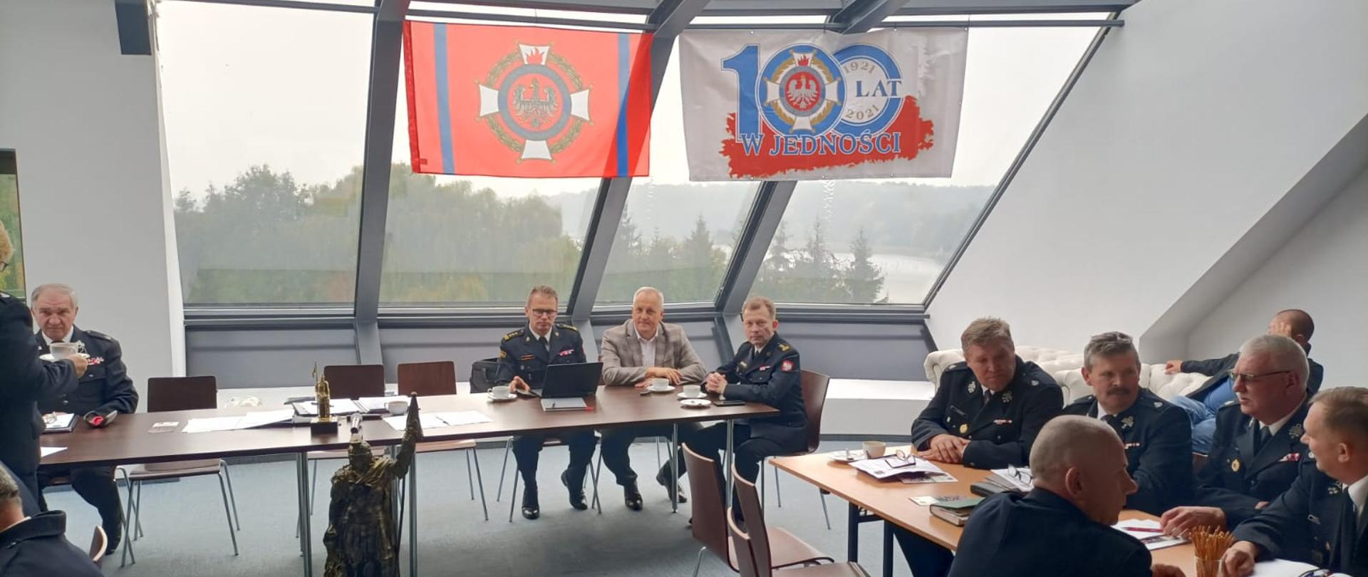 Zdjęcie wykonano w ciągu dnia, na przeszklonej sali delegaci Związku Ochotniczych Straży Pożarnych województwa kujawsko- pomorskiego ubrani w mundury galowe. W spotkaniu uczestniczy Zastępca Kujawsko-Pomorskiego Komendanta Wojewódzkiego PSP st. bryg. Marek Rusoń ubrany w mundur wyjściowy, starosta sępoleński Jarosław Tadych oraz Komendant Powiatowej Straży Pożarnej st. bryg. Maciej Suchomski również w mundurze wyjściowym.