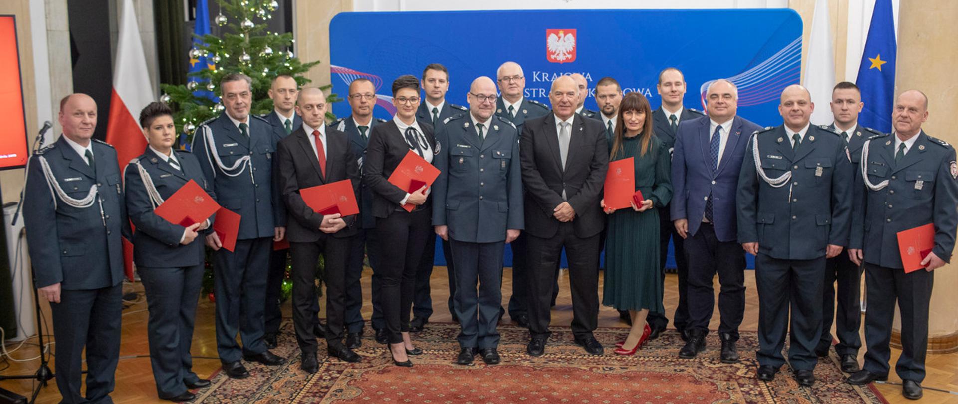 Minister finansów Tadeusz Kościński, Szef KAS Piotr Walczak oraz wyróżnieni pracownicy i funkcjonariusze Krajowej Administracji Skarbowej na tle baneru z godłem RP.