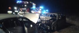 Dwa srebrne samochody osobowe po zderzeniu czołowym stojące naprzeciw siebie. Przody obu pojazdów zniszczone w dużym stopniu. Na jezdni i poboczu widoczne połamane elementy karoserii. w Tle strażacy i samochód ratowniczo-gaśniczy a za nim karetka pogotowia ratunkowego i widoczne błyskające na niebiesko światła pojazdów służb ratunkowych.
