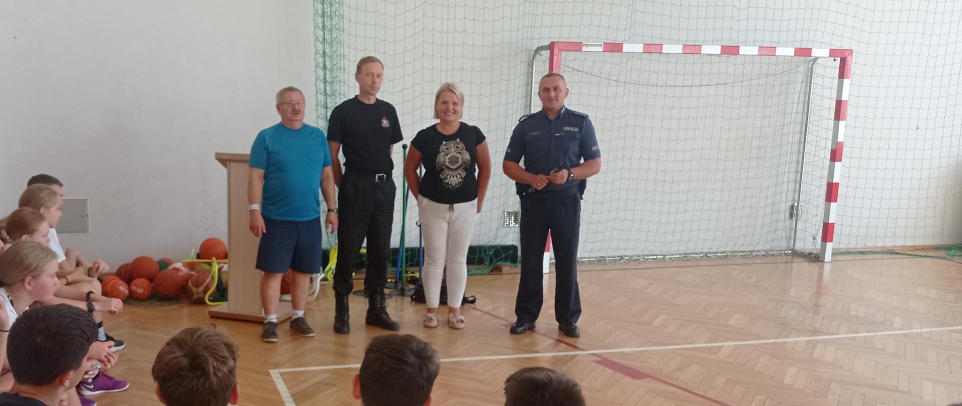 Zdjęcie przedstawia grupę młodzieży – uczestników obozu sportowego siedzących na podłodze wokół trojga prelegentów – przedstawiciela państwowej straży pożarnej, policji, sanepidu oraz kierownika grupy. Drugi plan zdjęcia stanowi bramka, tablica do koszykówki i piłki na tle wnętrza hali sportowej.