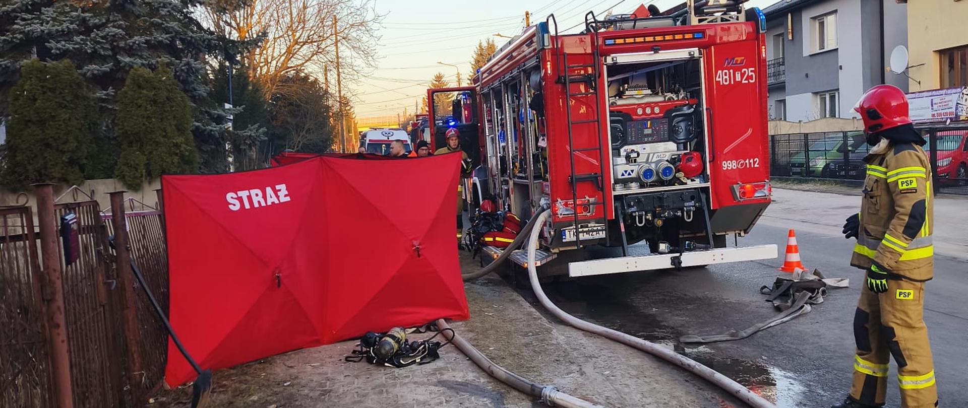 Na ulicy widać kilka wozów strażackich oraz strażaków prowadzących działania. Węże gaśnicze są rozwinięte na jezdni i podłączone do samochodów. Po lewej stronie ustawiono czerwony parawan zasłaniający część terenu. W tle znajdują się domy jednorodzinne i drzewa. Scena przedstawia szeroko zakrojoną akcję ratowniczo‑gaśniczą.