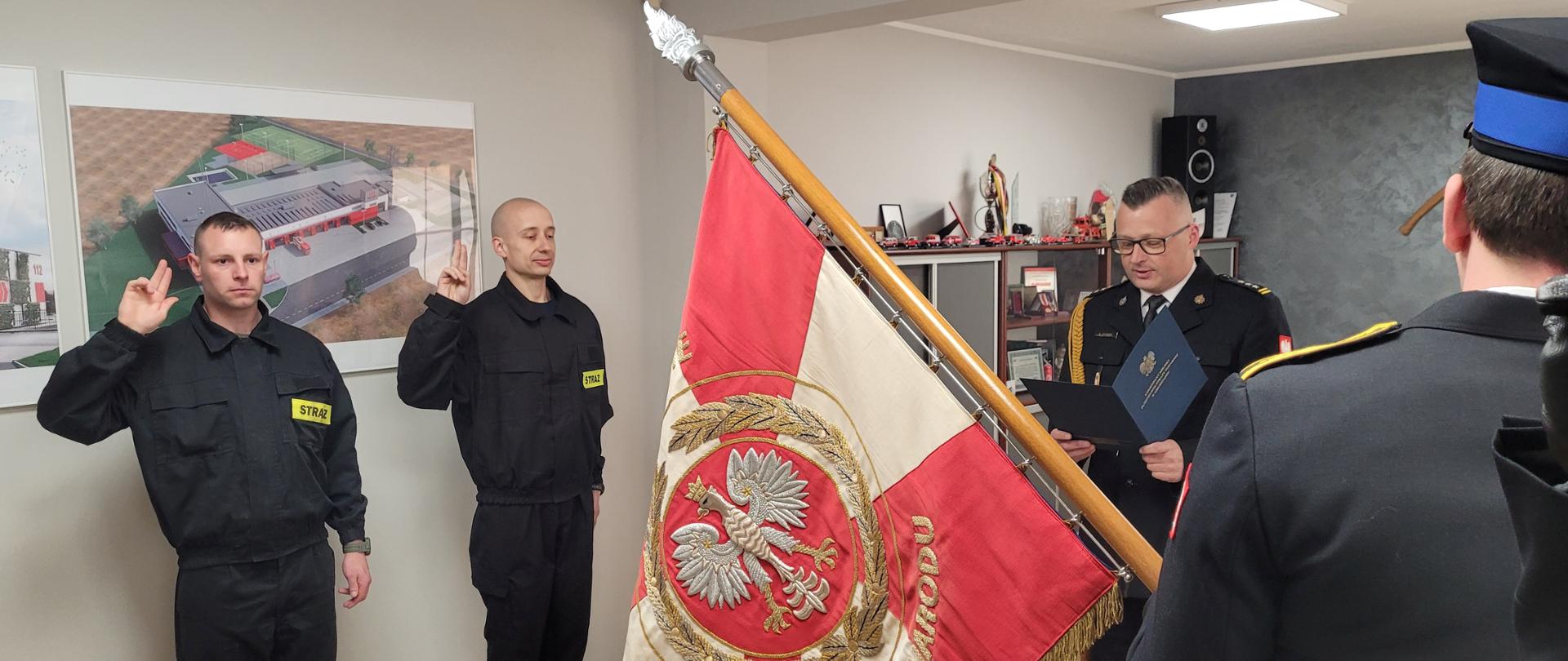 Gabinet, w którym zbiórkę maja strażacy. Dwóch stoi po lewej stronie i z podniesionymi prawymi rękoma składa ślubowanie. Jeden z teczką stoi na wprost.