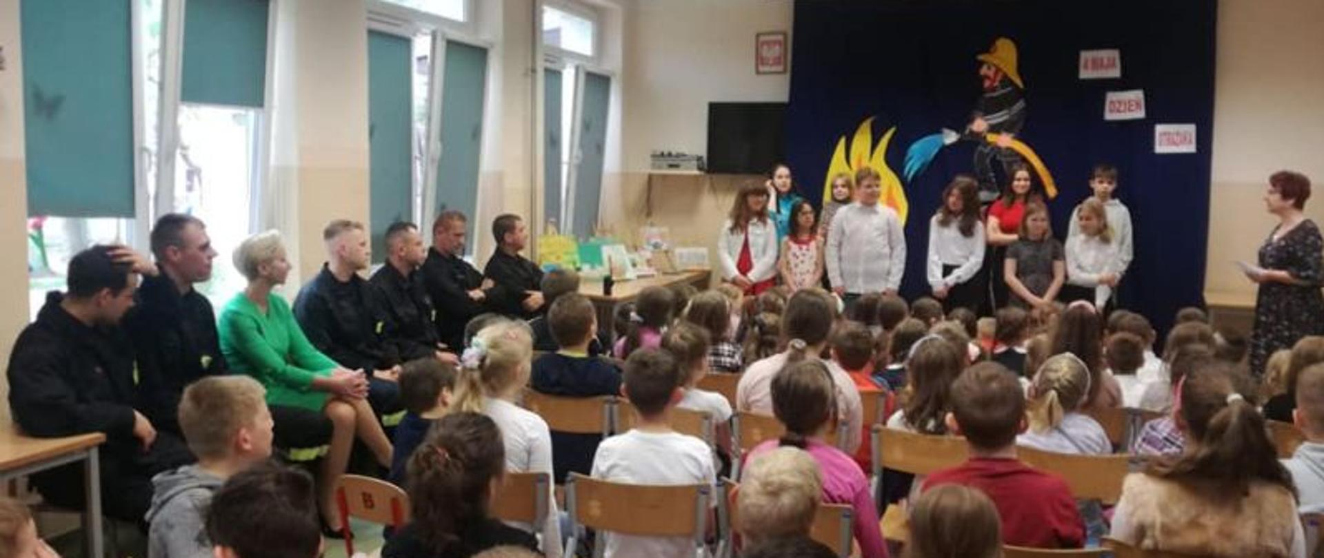 Zdjęcie wykonane wewnątrz szkoły. Widok na klasę szkolą wypełnioną dziećmi. Dzieci stoją również w oddali przy tablicy. Z lewej strony pod oknem siedzi zmiana służbowa wizytująca szkołę.