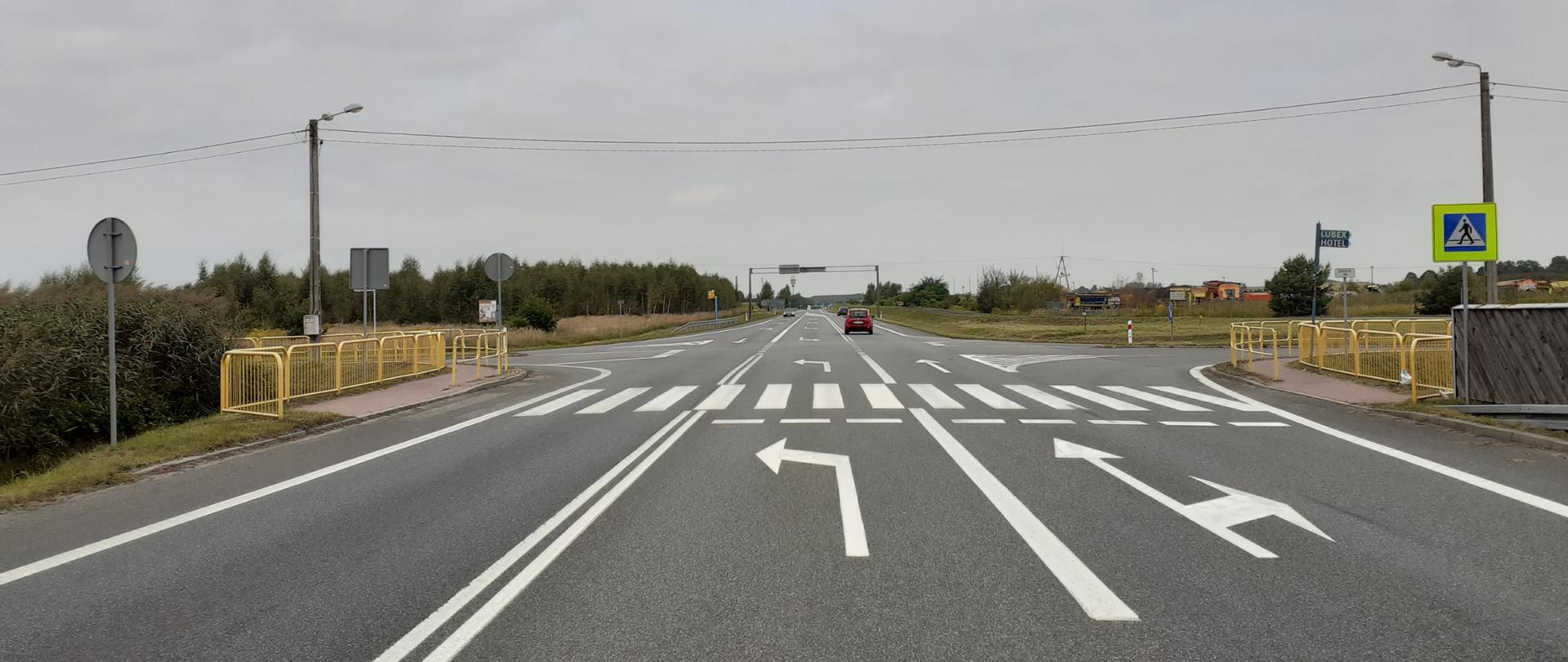 Fotografia przedstawia przejście dla pieszych przez 3-paswą drogę krajową nr 1 przed skrzyżowaniem w Lublińcu. Chodnikowe dojście do przejścia dla pieszych zabezpieczono barierami odgradzającymi w kolorze żółtym. Na drodze widoczne poruszające się pojazdy osobowe. Przejście oznakowane jest znakiem D-6 "przejście dla pieszych" na żółtym fluorescencyjnym tle. 