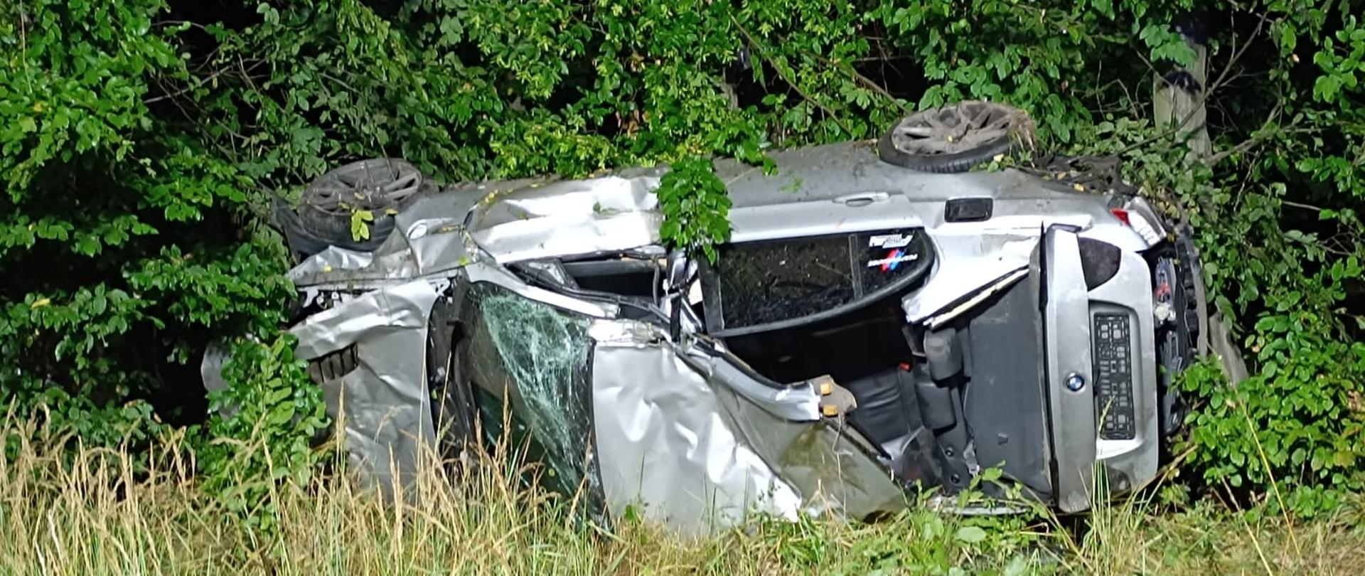 Samochód osobowy koloru srebrnego leży na lewym boku, w krzakach, widoczny zniszczony dach pojazdu