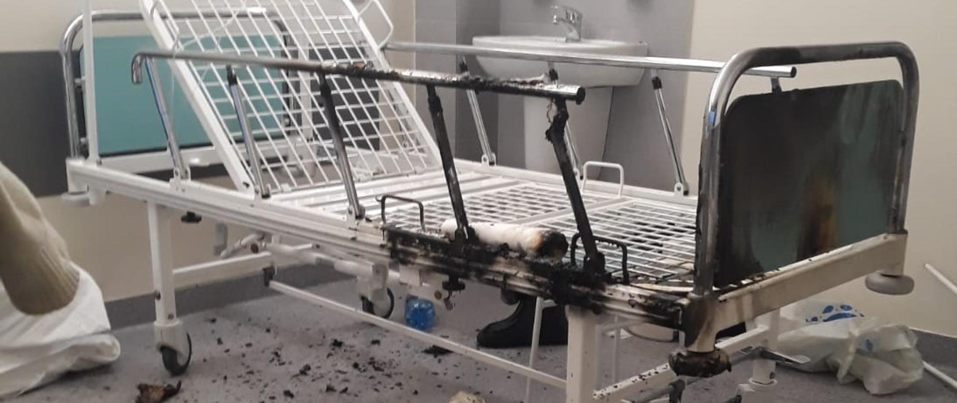 Na zdjęciu szpitalne łóżko. Fragment poręczy nadpalony. Nadpalone elementy na podłodze. Pomieszczenie szpitalne, oświetlenie sztuczne.
