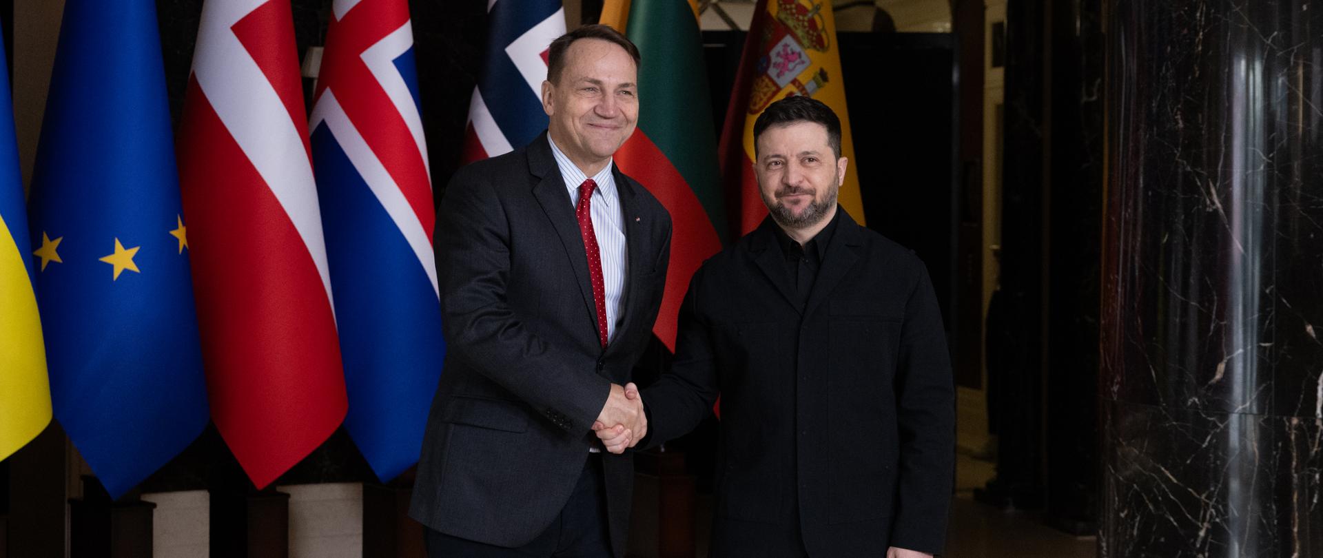 The photo shows two men in dark suits (Deputy Prime Minister and Minister of Foreign Affairs of Poland Radosław Sikorski and President of Ukraine Volodymyr Zelensky). They are shaking hands. Behind them are the flags of their countries. 