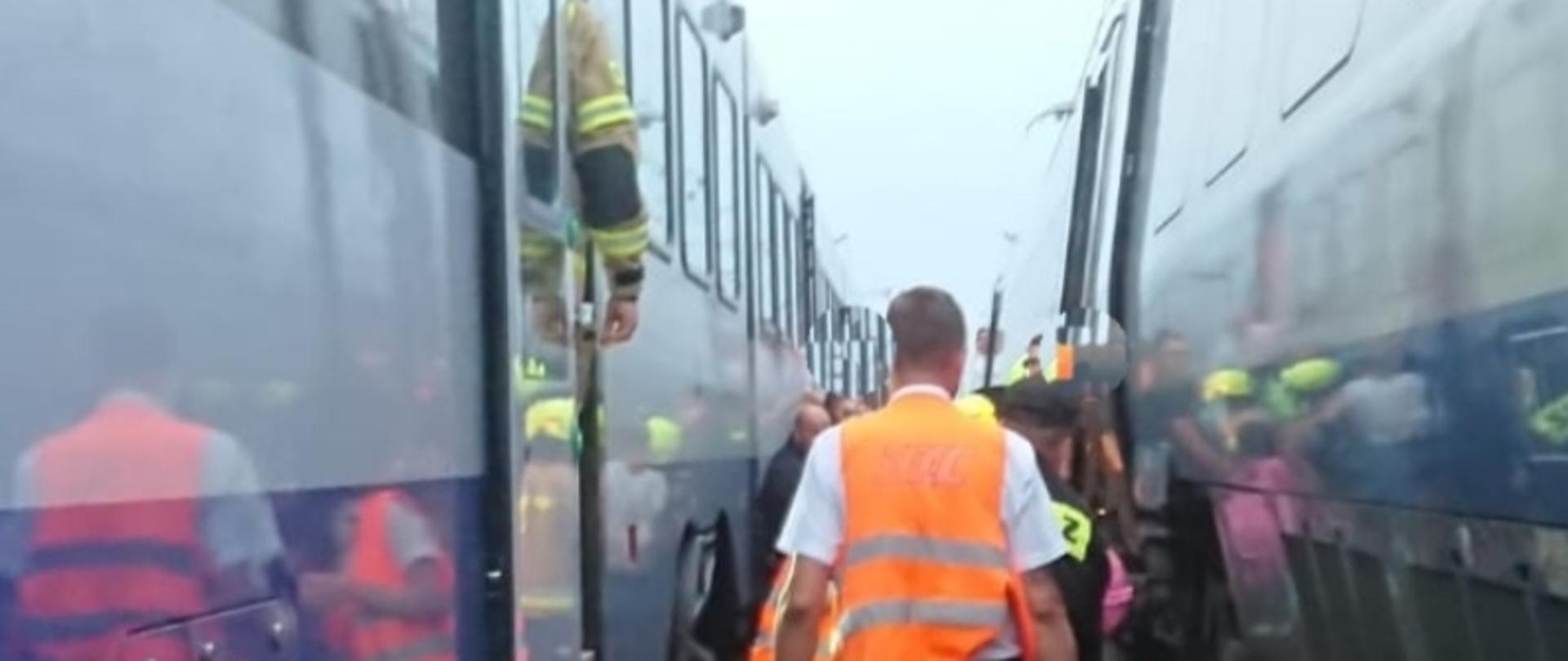 Szlak kolejowy, na dwóch sąsiednich torach dwa składy pociągu, pomiędzy nimi kierownik jednego ze składów na drugim planie przesiadający się pasażerowi, którym pomocy udzielają strażacy