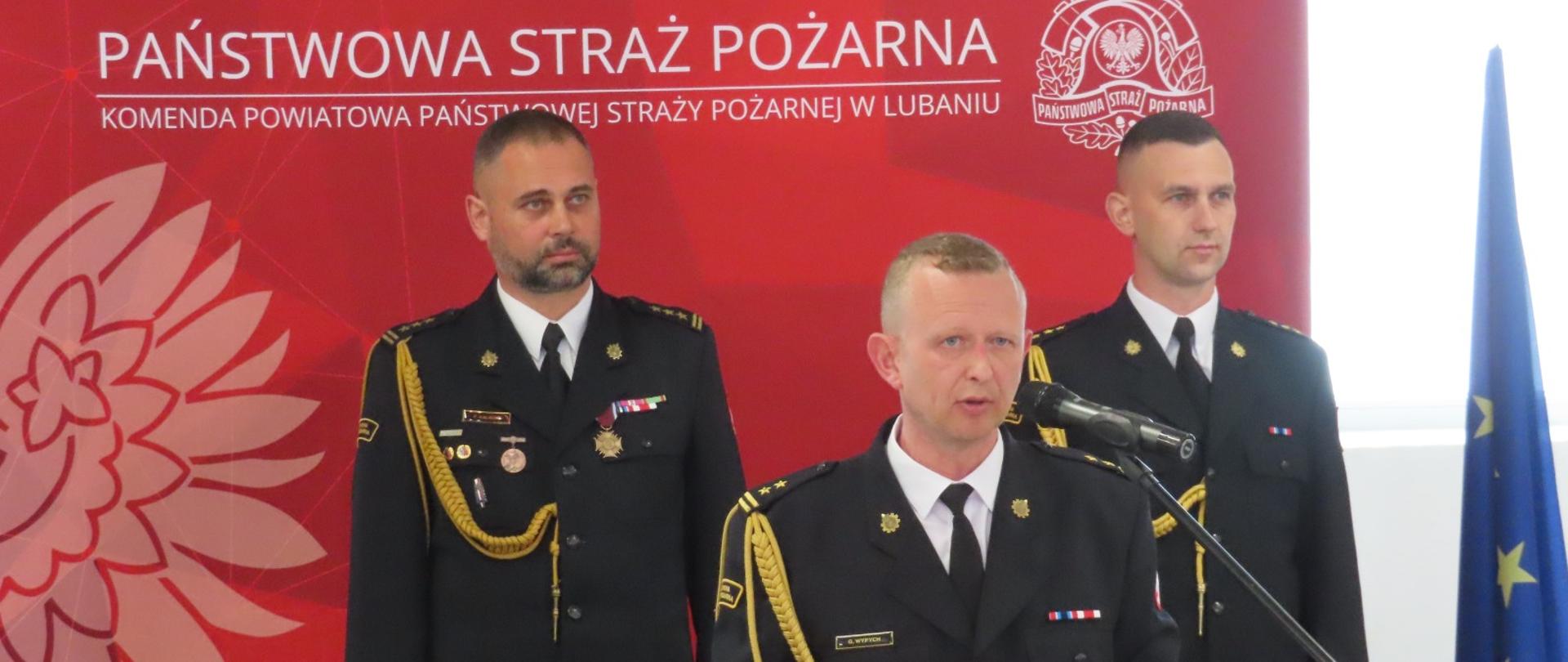 Dzień Strażaka w Komendzie Powiatowej PSP w Lubaniu
Komendant Powiatowy wygłasza przemówienie, za nim stoją zastępca Dolnośląskiego Komendanta Wojewódzkiego Państwowej Straży Pożarnej i zastępca Komendanta Powiatowego Państwowej Straży Pożarnej. W tle czerwony baner z białym logotypem Państwowej Straży Pożarnej i białym napisem Komenda Powiatowa Państwowej Straży Pożarnej w Lubaniu. Po prawej stronie znajduje się flaga Unii Europejskiej.