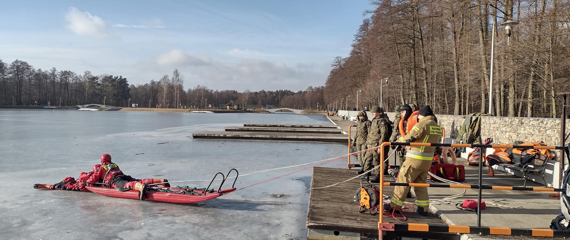 Na zdjęciu widać dwóch strażaków na lodzie. Jeden ze strażaków udaje osobę poszkodowaną, drugi zaś próbuję wydostać go przy pomocy sań lodowych na brzeg zbiornika wodnego. W tle uczestnicy szkolenia - żołnierze WOT