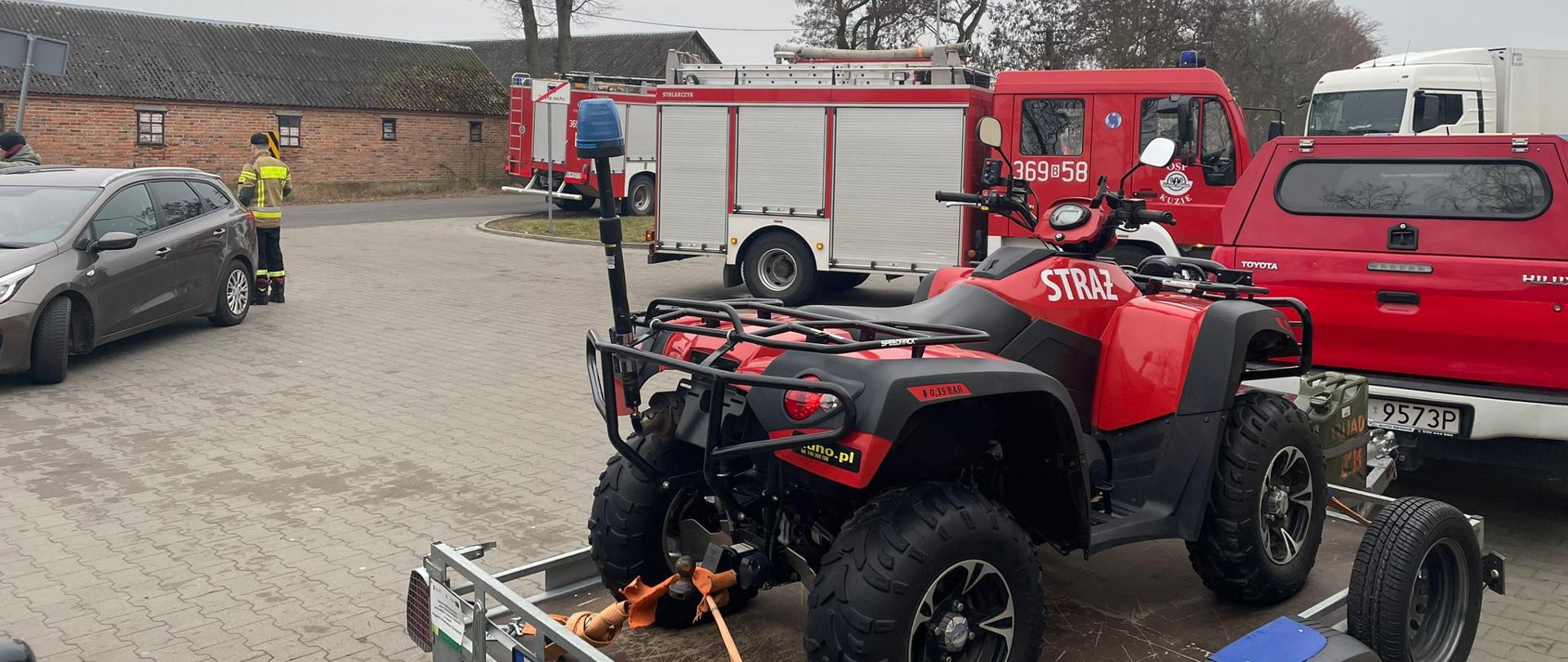 Na pierwszym planie na palcu stoi na przyczepce Quad, przed nim jest samochód terenowy oraz samochód ratowniczo-gaśniczy. Po lewej stronie jest samochód osobowy oraz obok niego stoi strażak. W tle za drogą stoi budynek gospodarczy, przed którym stoi samochód pożarniczy.