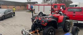 Na pierwszym planie na palcu stoi na przyczepce Quad, przed nim jest samochód terenowy oraz samochód ratowniczo-gaśniczy. Po lewej stronie jest samochód osobowy oraz obok niego stoi strażak. W tle za drogą stoi budynek gospodarczy, przed którym stoi samochód pożarniczy.