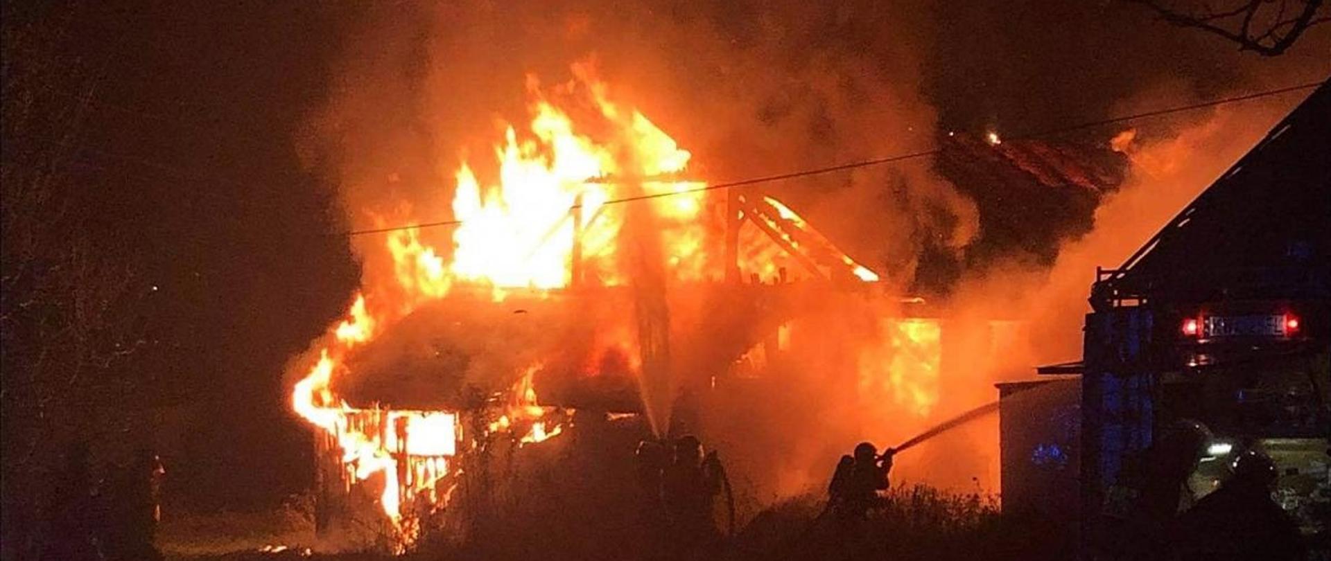Na środku budynek objęty pożarem w nocy. Płomienie wychodzą przez przepalone ściany. Przed budynkiem strażacy. W środkowej części trzech strażaków trzyma linie wężowe i dwa strumienie wody na płonący budynek. Po prawej stronie tył samochodu pożarniczego i dwóch strażaków zajętych obsługą sprzętu.