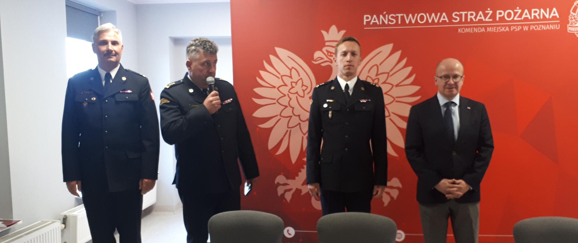 czterech mężczyzn stoi w szeregu, trzech z nich to strażacy, jeden w garniturze, w tle czerwony baner