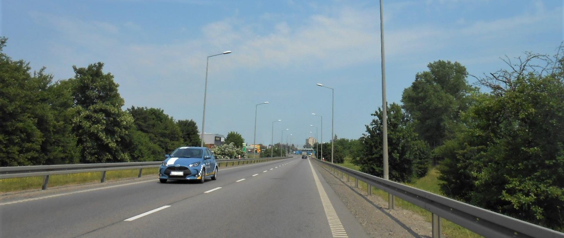 Zdjęcie przedstawia drogę krajową nr 91 w Tczewie. Na pierwszym planie jest droga z pojazdami, ale brak jest na niej chodnika i ścieżki rowerowej. Po obu stronach drogi są tereny zielone z drzewami.