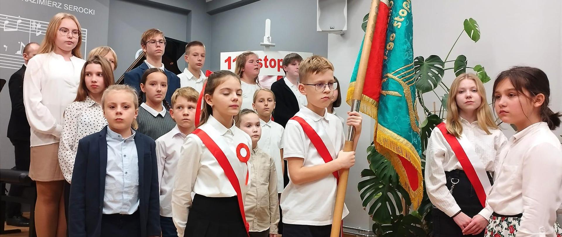 Zdjecie przedstawia uczniów szkoły muzycznej podczas uroczystości Dnia Niepodległości, uczniowie ubrani na galowo stojących na scenie w auli szkoły