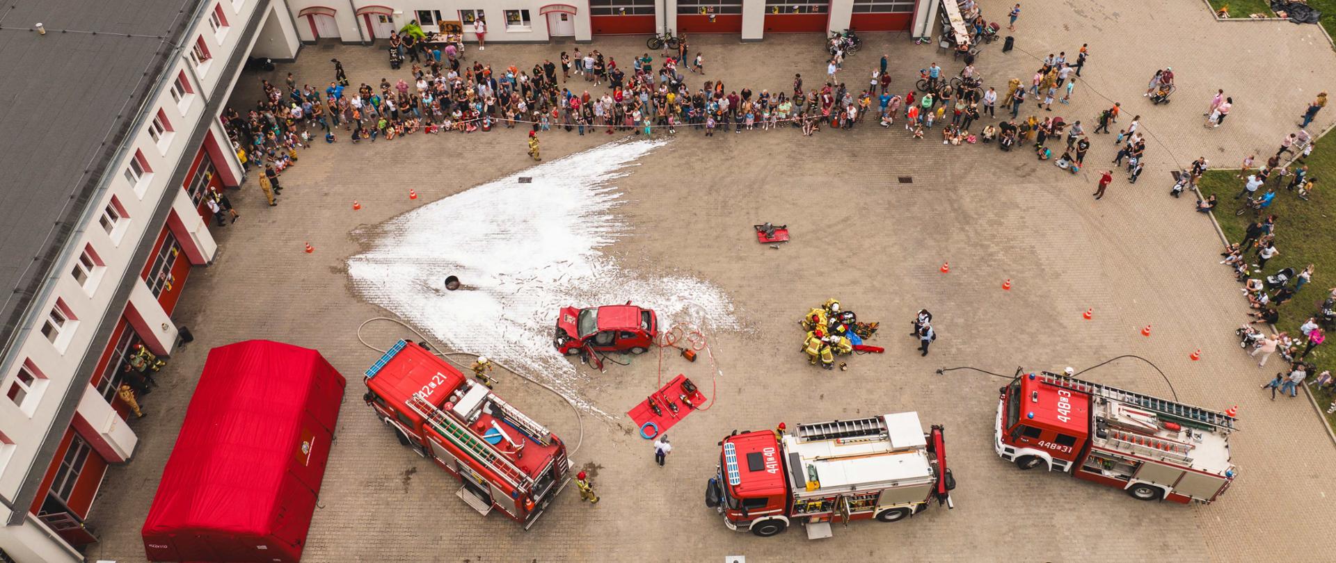 Widok z lotu ptaka utwardzonego miedzy budynkami placu, na którym stoją 3 strażackie samochody a ich załogi prowadzą symulowaną akcję ratowniczą. Pokaz ogląda zgromadzona publiczność.