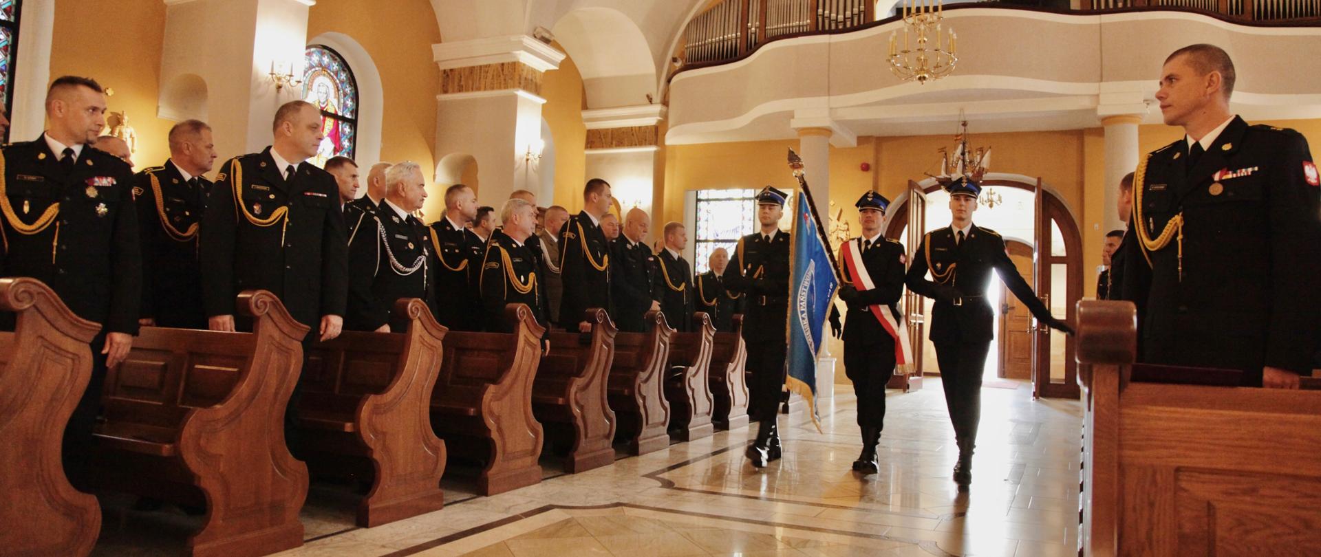 Strażacy stoją w ławkach w kościele. Wszyscy są ubrani w mundury galowe. Przez środek kościoła wchodzi poczet sztandarowy składający się z trzech strażaków. Środkowy z nich niesie sztandar oraz ma przepaskę w kolorach białym i czerwonym.