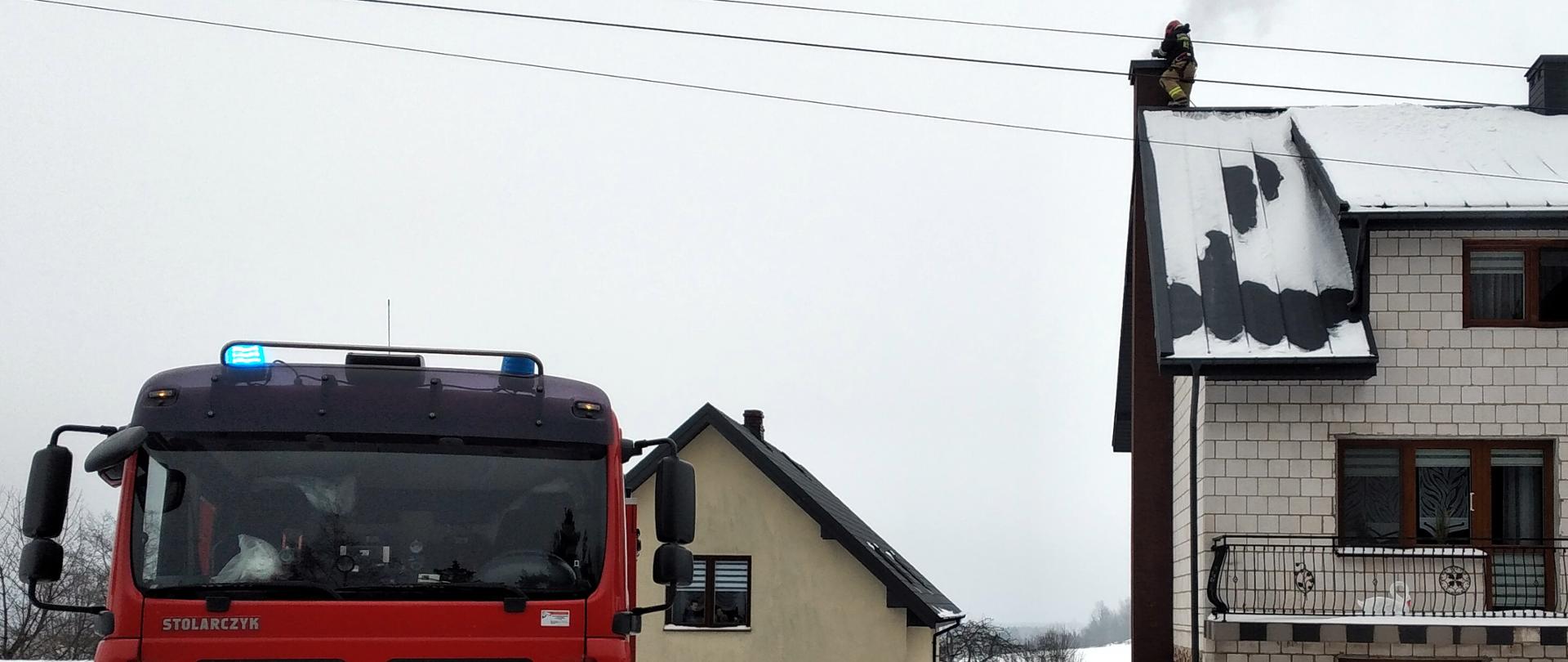 Zdjęcie przedstawia budynek jednorodzinny w miejscowości Malmurzyn z samochodem pożarniczym z JRG nr 2 w Kielcach na pierwszym planie. W oddali na dachu budynku przy kominie znajduje się strażak. Z komina wydobywa się lekki dym.