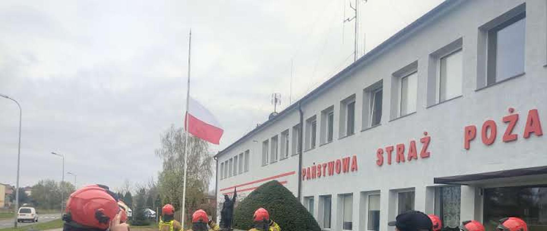Zdjęcie przedstawia uroczystą zmianę służby przed budynkiem Komendy Powiatowej Państwowej Straży Pożarnej w Świdwinie. Trzech strażaków pod masztem flagowym oddaje honory do Flagi Państwowej. Przed asystą honorową znajdują się w szeregu równolegle do siebie zwrócone zmiany.
