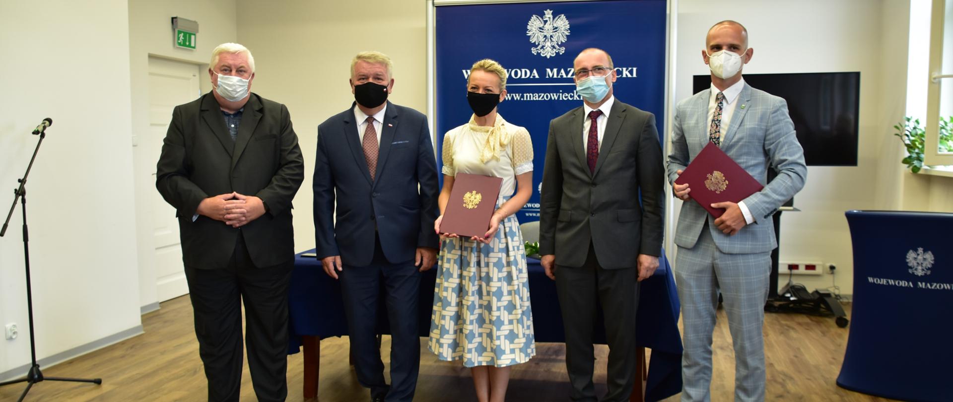 Na zdjęciu znajduje się pięć elegancko ubranych osób - kobieta i czterech mężczyzn. Pozują oni na granatowej ściance wizerunkowej Wojewody Mazowieckiego. Niektórzy z nich trzymają w dłoniach umowę na realizację zadań w ramach Rządowego Funduszu Rozwoju Dróg. 