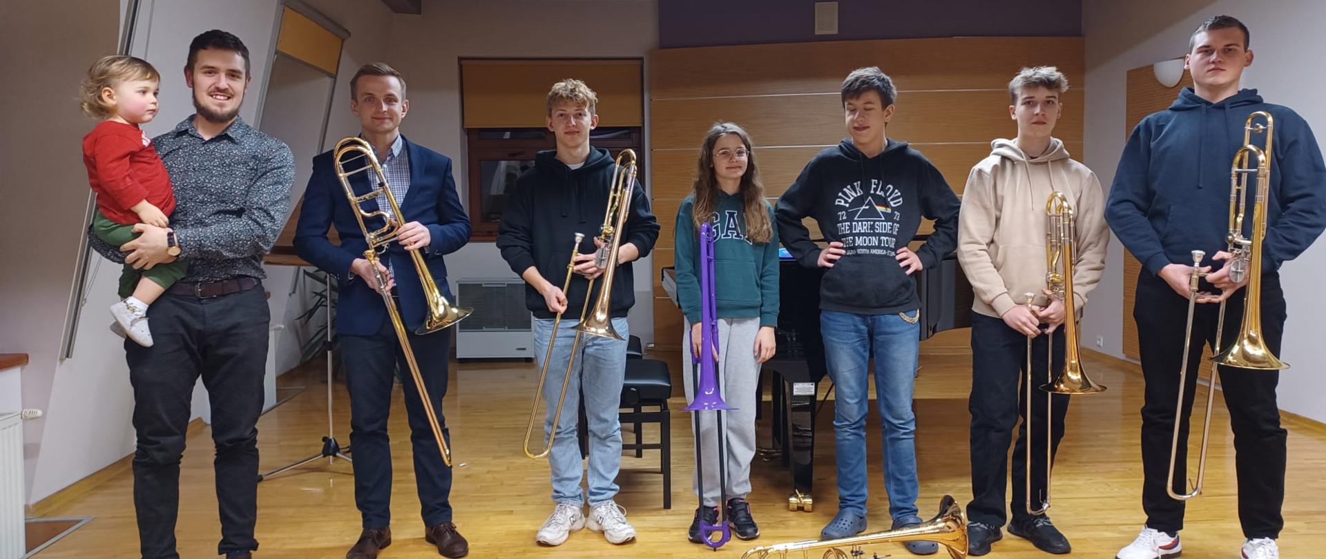 Zdjęcie grupowe. Uczniowie, prowadzący warsztaty i nauczyciel puzonu stoją w jednym rzędzie na scenie sali kameralnej i trzymają w dłoniach puzony. Za nimi dwa fortepiany oraz dwie ławy fortepianowe. Trzy okna przysłonięte roletami. Pod oknami kaloryfery a przy jednym z nich nawilżacz. Nad nimi zapalone przy suficie zapalona lampa