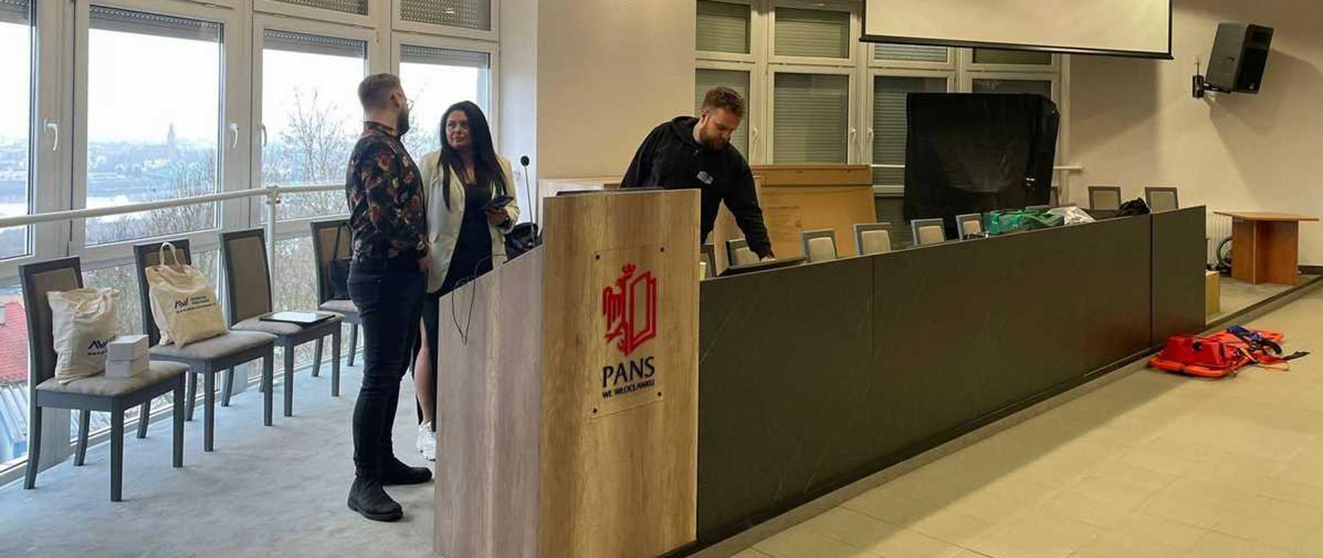 Na zdjęciu widać salę konferencyjną lub wykładową, w której przygotowywane jest wydarzenie. Na mównicy znajduje się logo PANS (Państwowej Akademii Nauk Stosowanych), co wskazuje, że miejsce to jest związane z tą uczelnią wyższą.