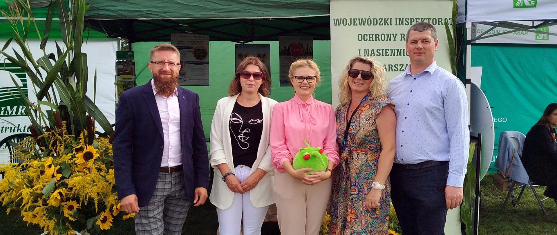 Grupa pięciu osób stoi przed zielonym namiotem wystawowym Wojewódzkiego Inspektoratu Ochrony Roślin i Nasiennictwa w Warszawie. Osoby uśmiechają się do zdjęcia, a jedna z nich trzyma pluszową maskotkę w kształcie zielonego owada. W tle widoczne są banery i dekoracje z kwiatów słoneczników.
