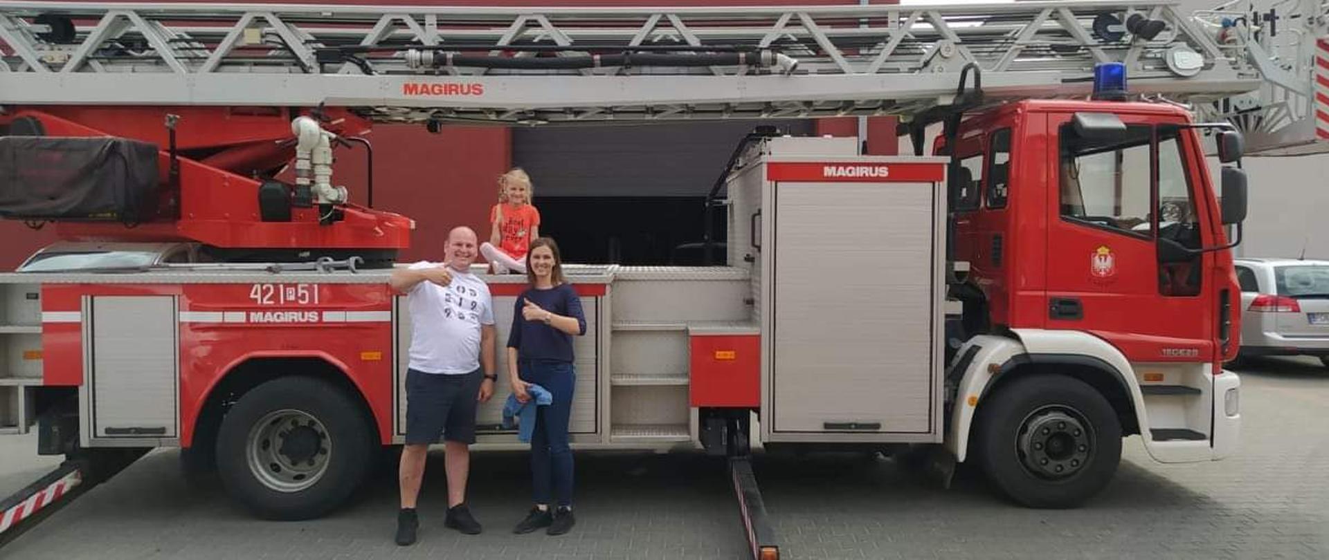 Dwie dorosłe osoby tj. mężczyzna w białej koszulce i granatowych spodenkach oraz kobieta w granatowych spodniach i bluzce stoją uśmiechnięci przed czerwonym samochodem strażackim. Prawe ręce w geście OK. Dziewczynka w pomarańczowej bluzce siedzi na podeście samochodu. W tle czerwony budynek warsztatowy.