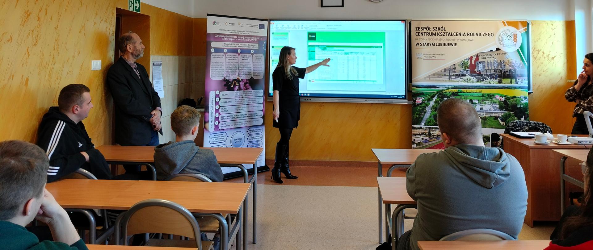 Młodzież siedzi w ławkach patrząc na prowadzącą szkolenie, która pokazuje coś na ekranie