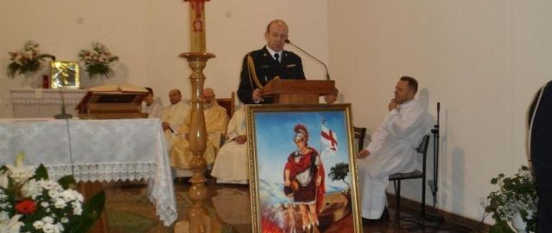 umundurowany strażak stojący przy mikrofonie na ołtarzu w kościele, przed nim stojący obraz św. Floriana, po lewej strona zapalona świeca
